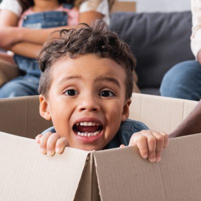 thrilled african american boy sitting in carton box near blurred family, banner